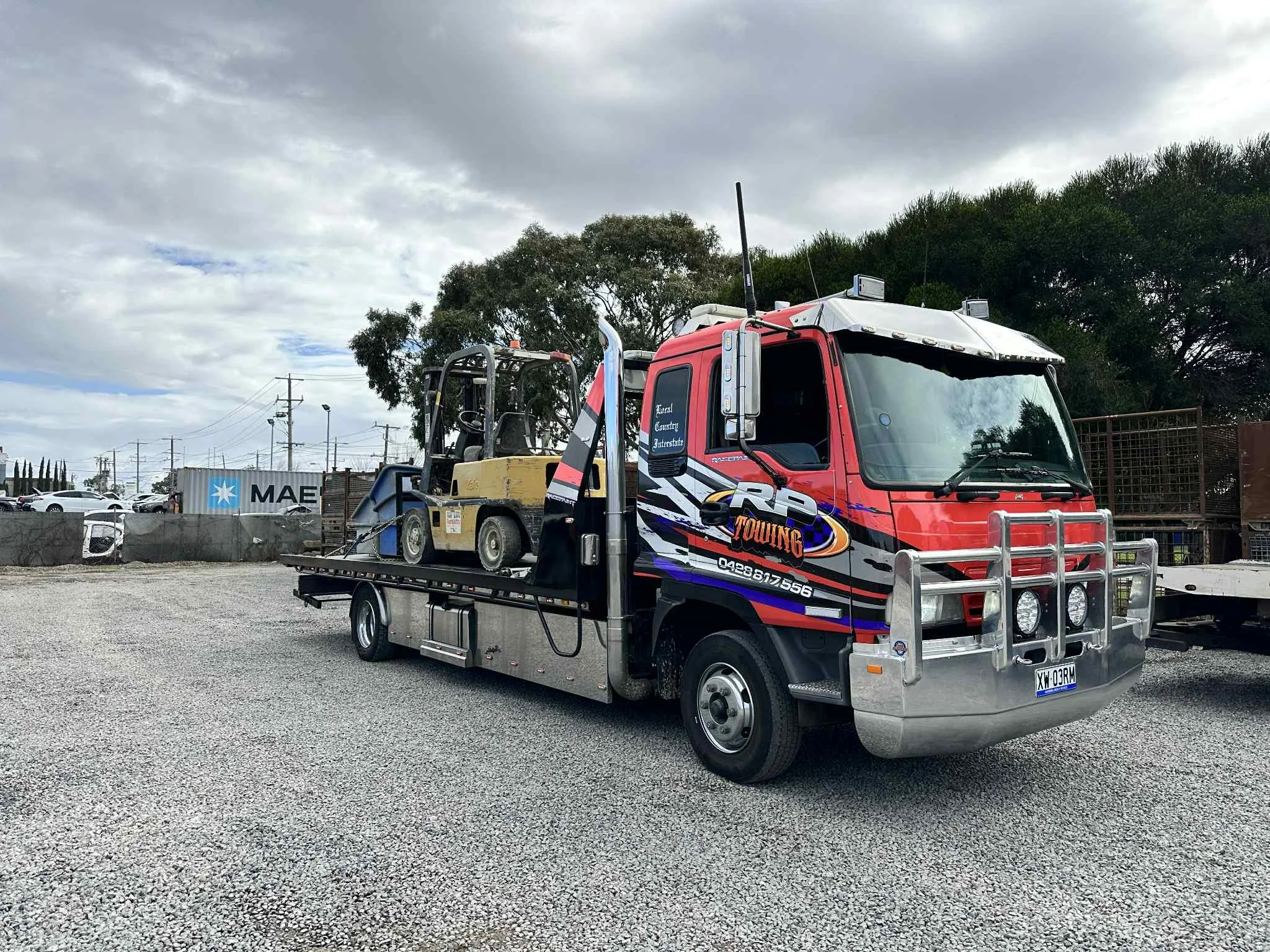A tow truck parked outdoors on a gravel surface, carrying a small yellow loader machine. The truck is black, red, and silver with towing graphics, and has a front bumper guard with four lights. The background shows cloudy skies, trees, power lines, a