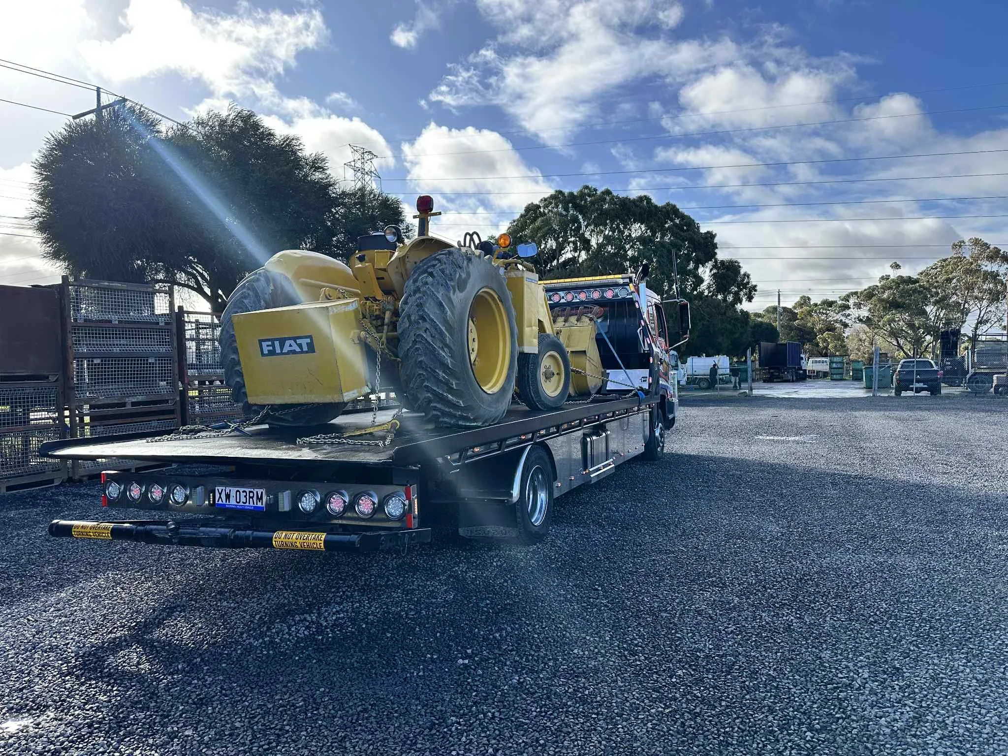 A flatbed tow truck carrying a small yellow Fiat vehicle with large tires and a chained down sign on a gravel lot.