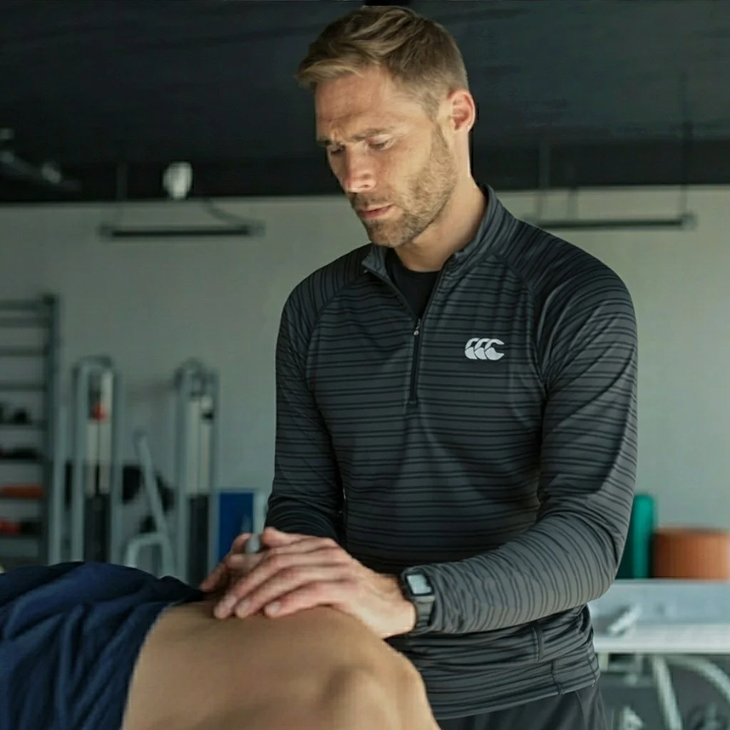 A male professional physio performing a physical examination on a patient's knee in a clinical setting.