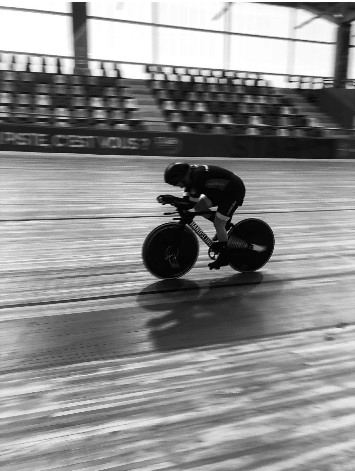 A cyclist wearing a helmet and sports gear riding a track bike on an indoor velodrome track with blurred background and empty seats.