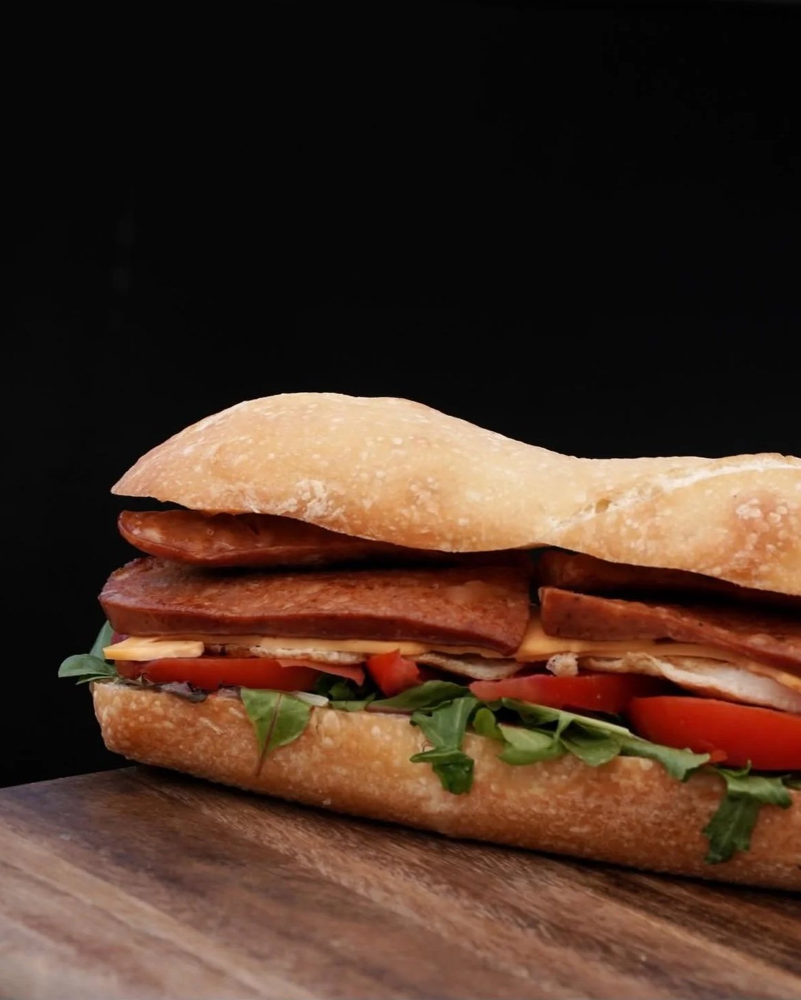 Submarine sandwich with bread, turkey or chicken, tomatoes, lettuce, and possibly bacon or ham, on a wooden surface against a black background.  Food Photo Co.