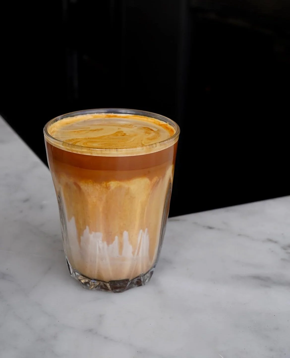 A glass of layered coffee beverage with a creamy, caramel-colored top and a lighter, milk-like bottom on a marble surface with a dark background.
