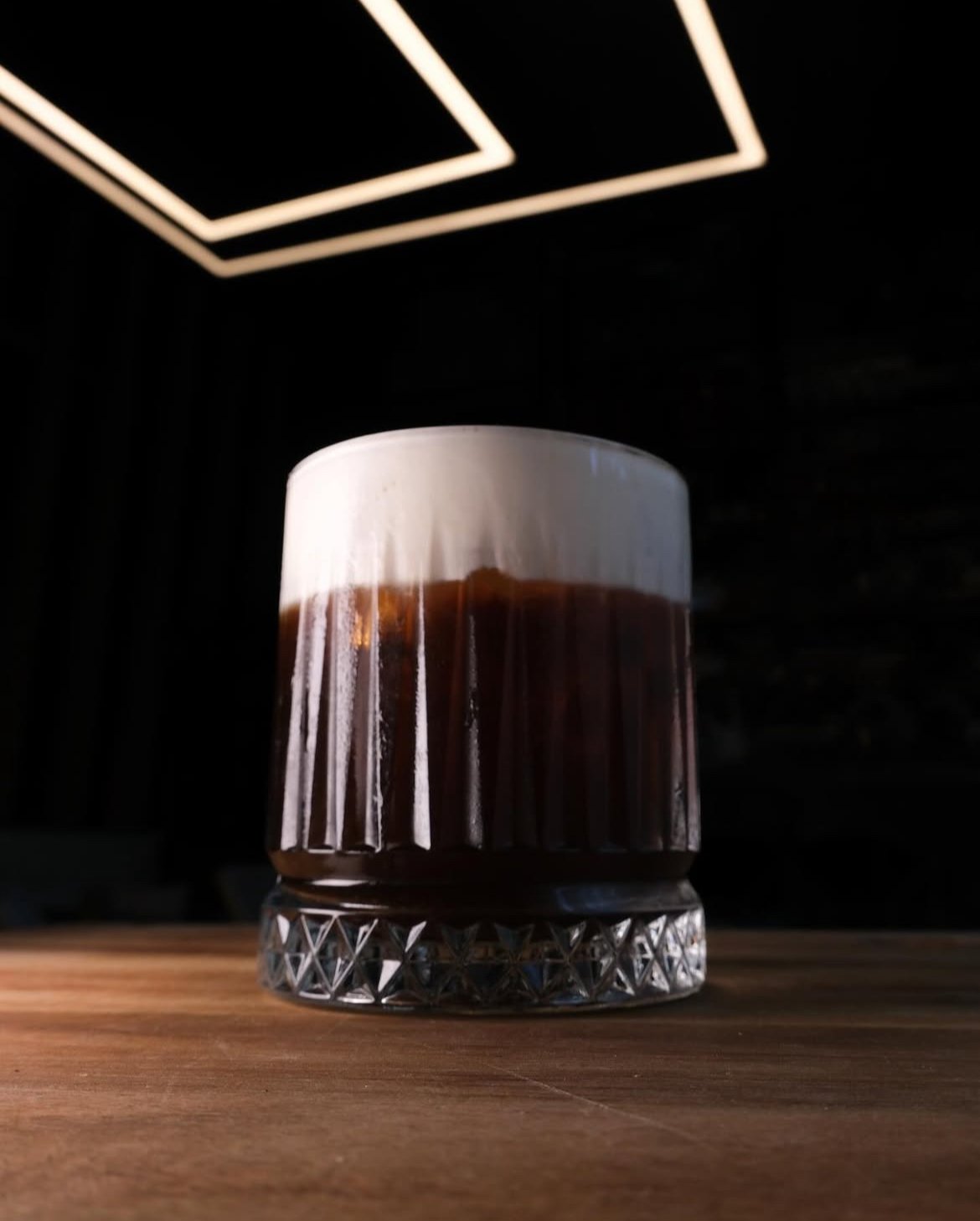 A glass of iced coffee with a thick layer of foam on top, placed on a wooden surface with a dark background and geometric ceiling lights.  Food Photo Co.