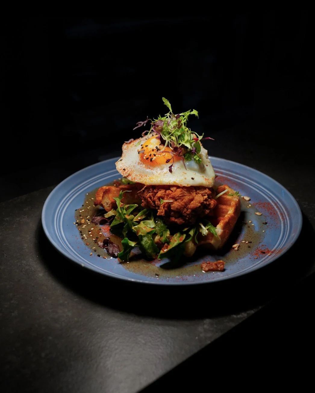 A blue plate of crispy fried chicken topped with a fried egg, garnished with microgreens, on a bed of salad greens, with waffle fries in the background, garnished with seasoning and sauce.