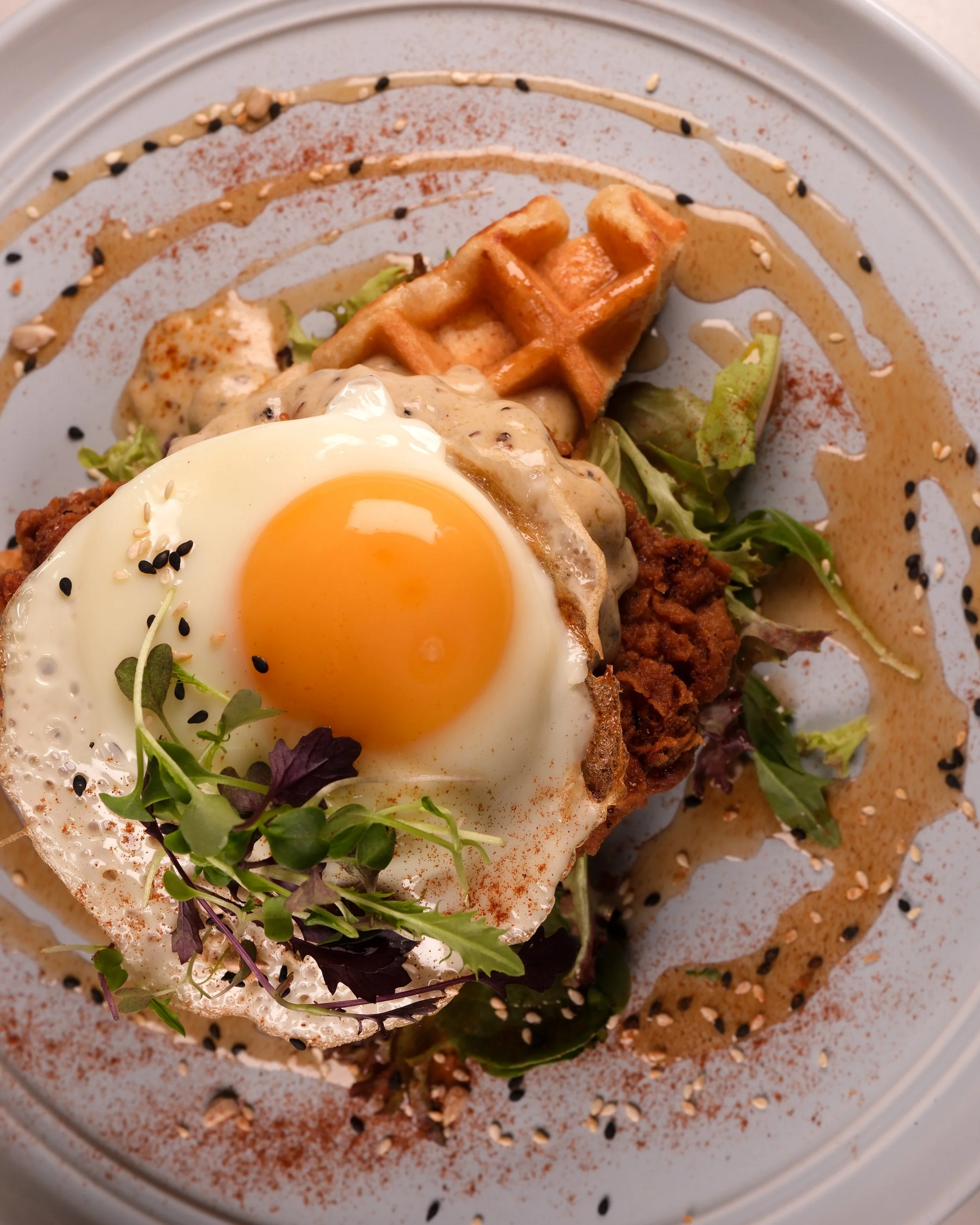 A breakfast plate with a fried egg topped with microgreens, a waffle, salad greens, a biscuit with gravy, and sauces decorated with black and white sesame seeds.  Food Photo Co.