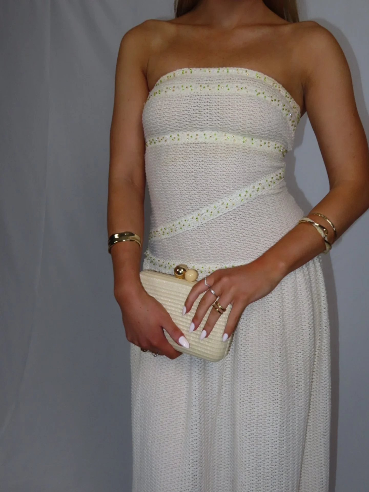 Woman wearing a strapless beige dress with a textured fabric, gold jewelry, holding a cream-colored clutch with decorative spheres.