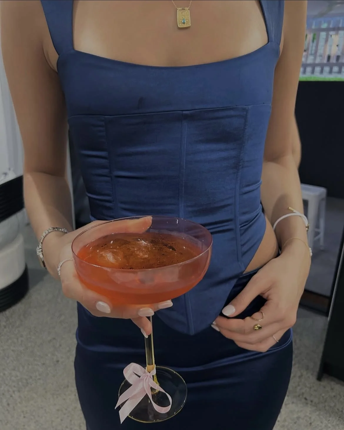 A woman in a blue dress holds a pink cocktail glass with a drink, wearing jewelry, with a background indicating an indoor setting.