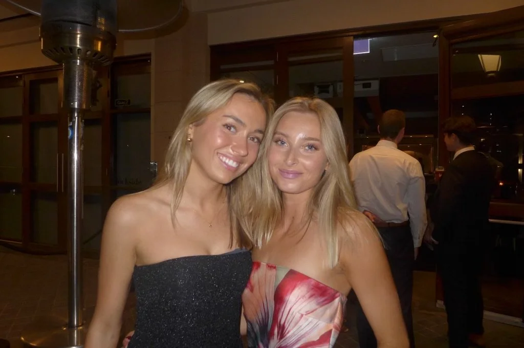 Two young women with blonde hair smiling and posing together indoors at a social event, with other people in the background.