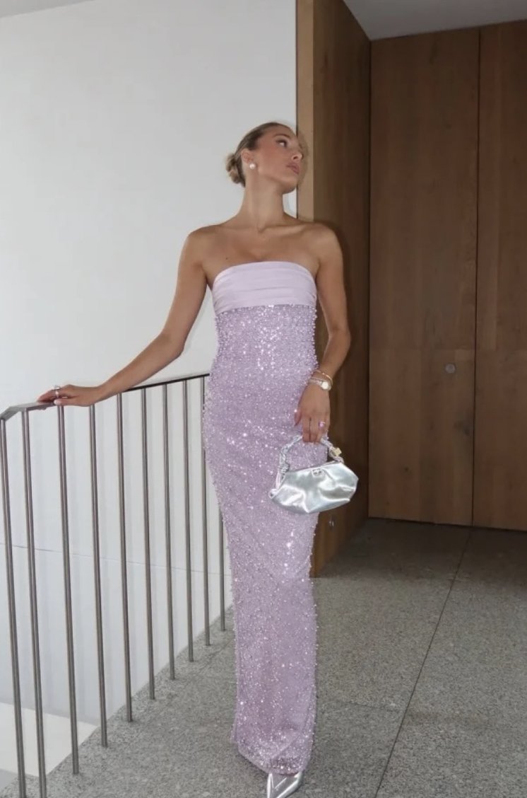 A woman in a strapless, floor-length, sequined pink gown standing indoors near a wooden wall and a staircase railing, holding a silver clutch purse, wearing a watch and earrings, with her hair styled in an updo.