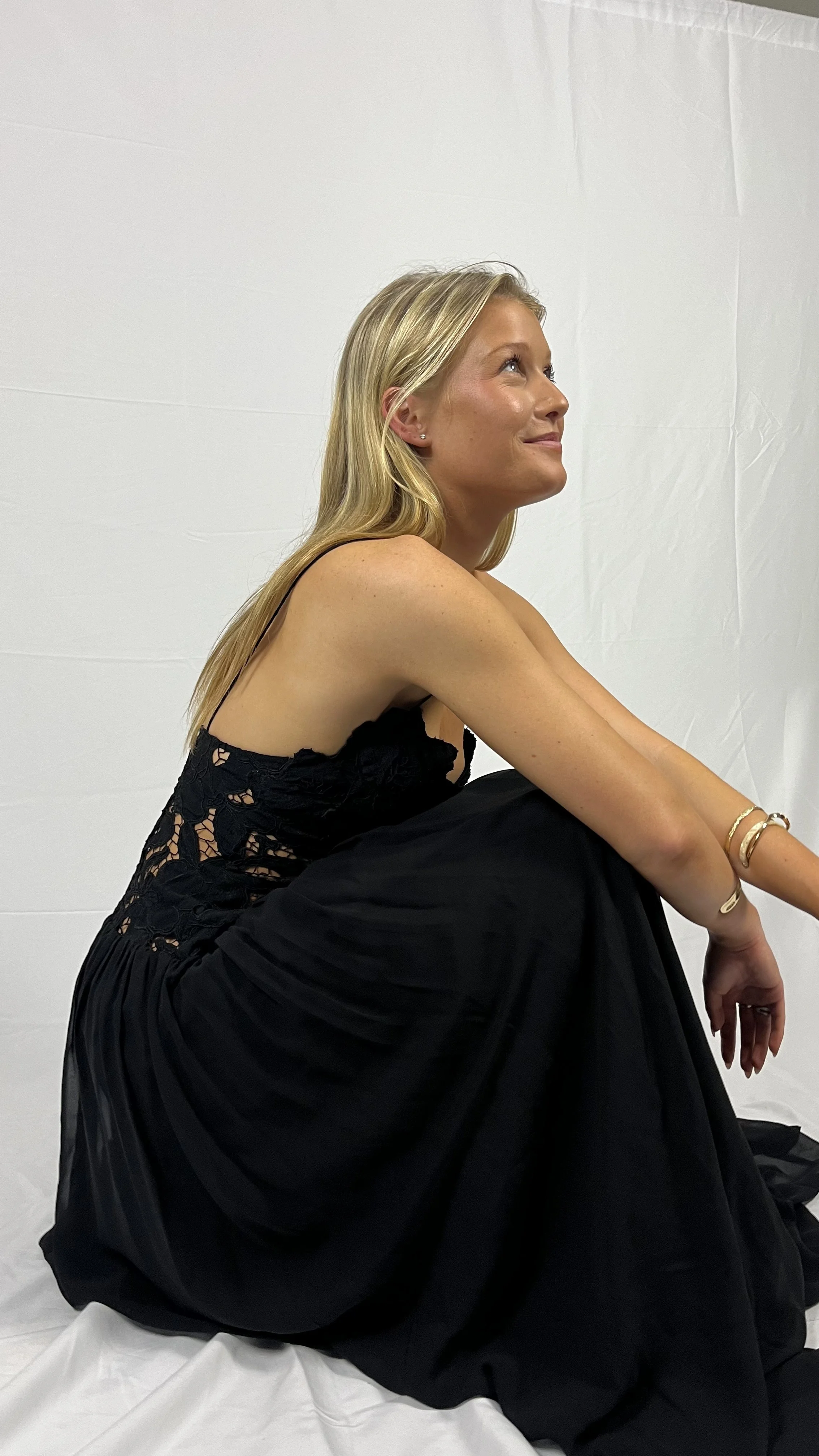 A woman sitting on the floor against a plain white background, wearing a black lace and pleated dress, with blonde hair and bracelets on her arm, looking upwards and smiling.