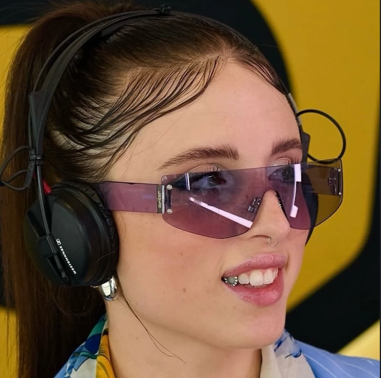 Una donna con acconciatura liscia porta occhiali da sole rosa e cuffie nere, sorridendo leggermente e con uno sfondo giallo e nero.
