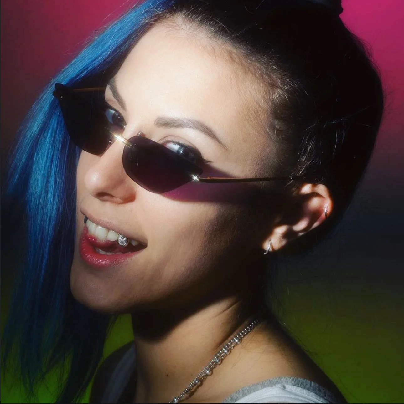 Ragazza con capelli corti neri e ciuffo blu, indossa occhiali da sole neri, ha un piercing sulla lingua e una collana, sfondo colorato con tono rosso e verde.