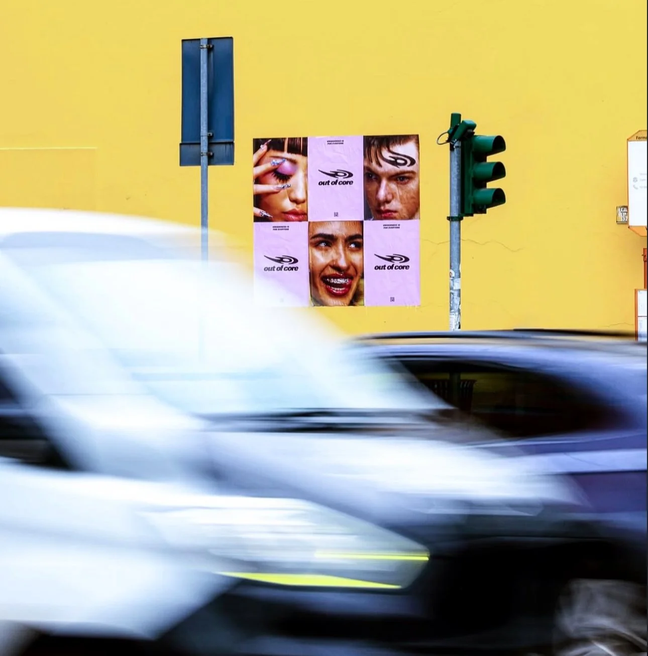 Immagine di un muro giallo con manifesti pubblicitari e semafori, con due auto in movimento davanti.