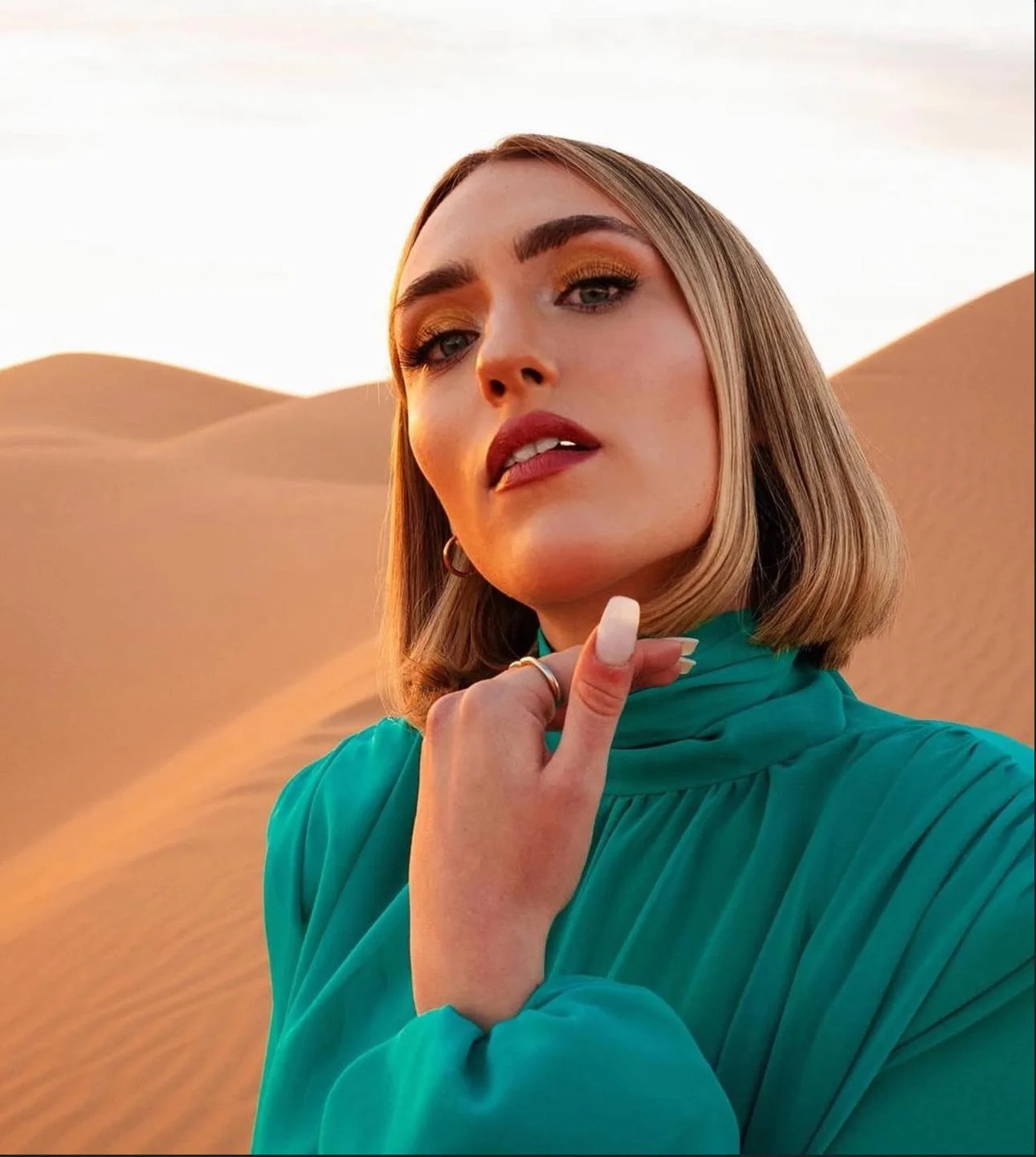 Una giovane donna con capelli corti in un deserto durante il tramonto, indossa un abito verde e ha un trucco marcato con ombretto dorato.