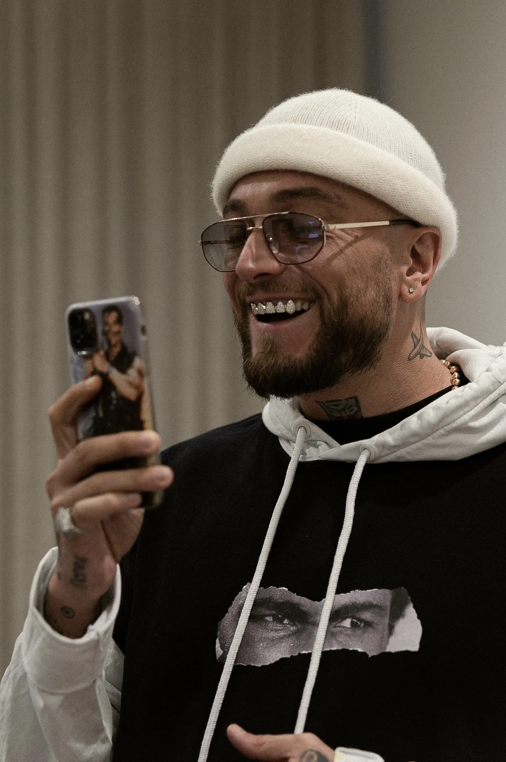Uomo sorridente con cappello bianco, occhiali da sole e tatuaggi, che guarda uno smartphone con una foto di un'altra persona sulla cover, in un ambiente interno.