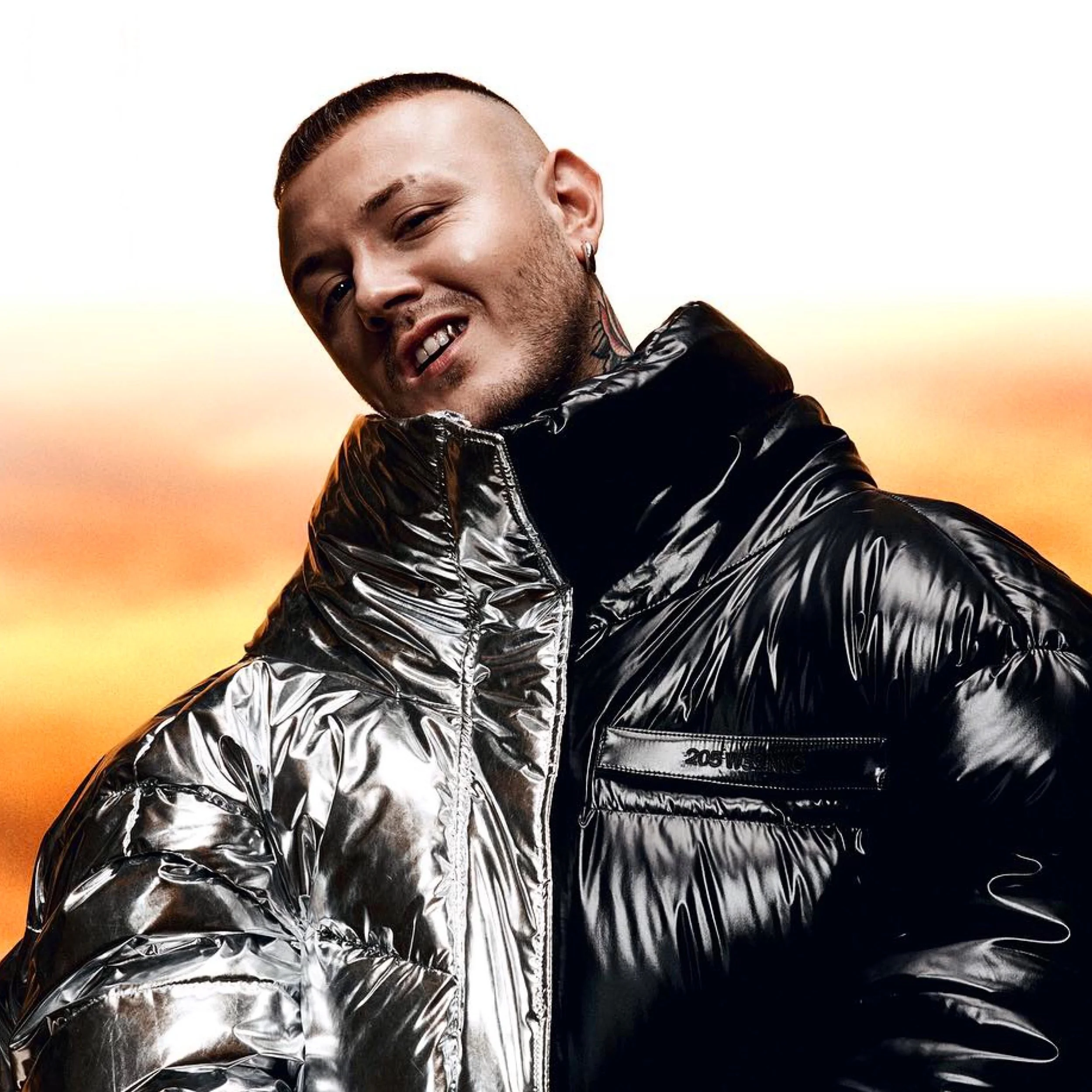 Uomo con capelli rasati, tatuaggi e orecchino, indossa una giacca nera brillante con collare alto, sfondo sfumato di colori caldi.