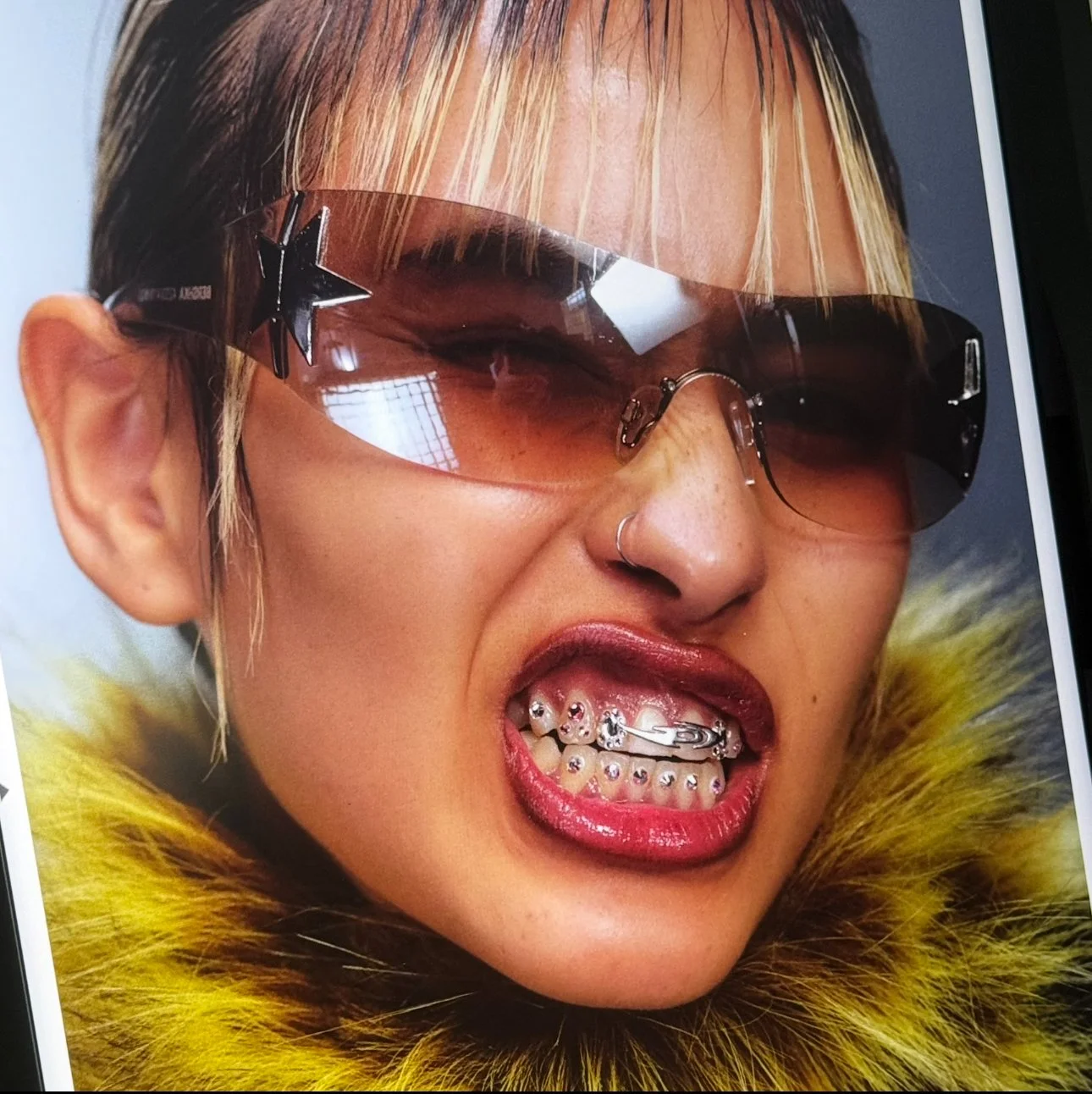 Una donna con occhiali da sole grandi e colorati, bocca aperta mostrando denti con piercing e un sorriso scorbutico, indossa un cortissimo cappotto di pelliccia gialla.