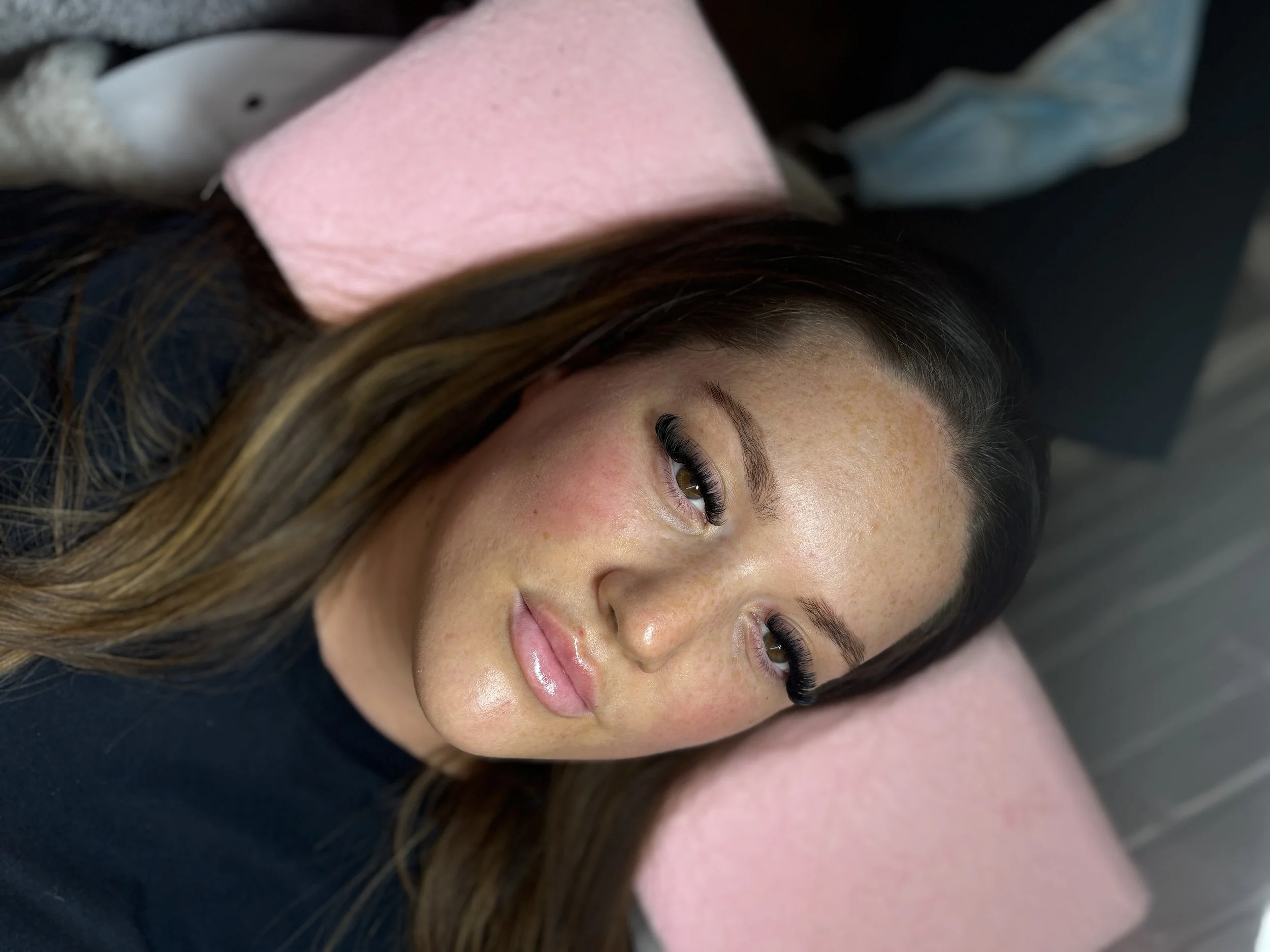 A woman with long brown hair lying on a pink pillow, looking at the camera with a slight smile, black top, and natural makeup showing freckles.