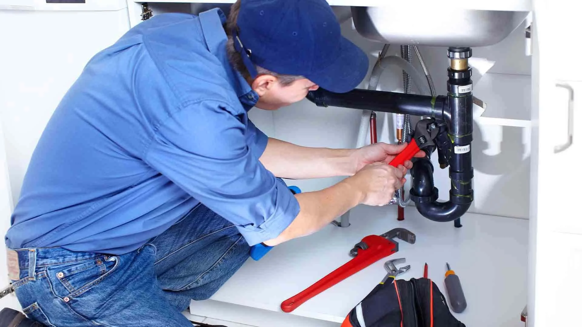 A man wearing a blue shirt and cap is working under a kitchen sink, using pliers to tighten or loosen a pipe fitting. Various tools and a toolbox are on the floor.