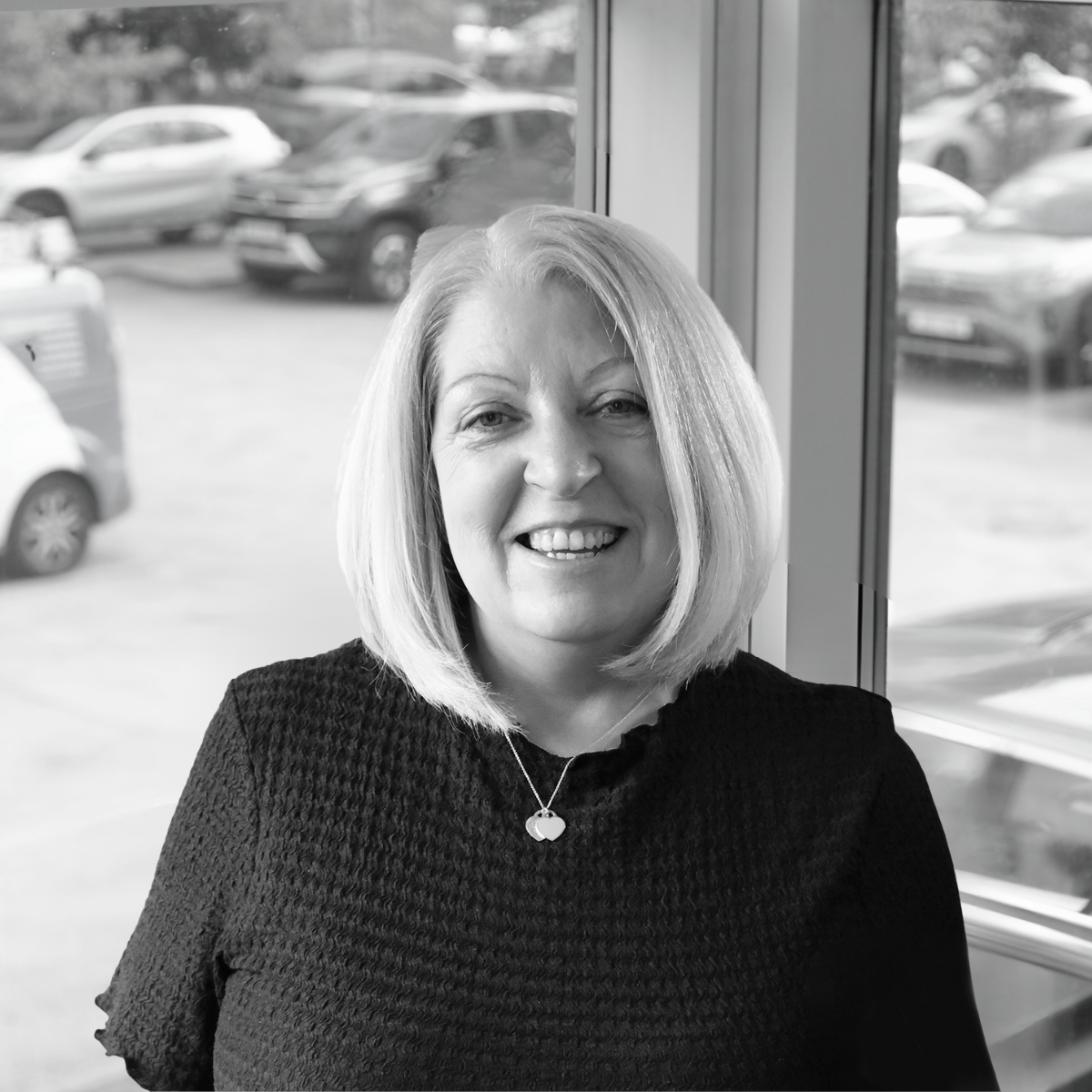 Shirley Renham - Black and white photo of a man in a suit jacket and white shirt, smiling and standing in front of a window with cars outside.
