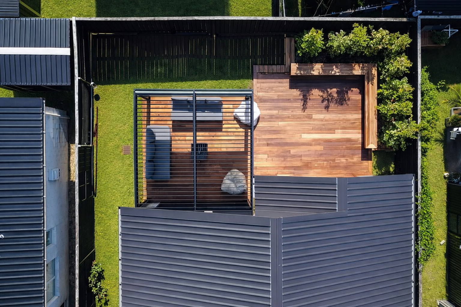 Bird's-eye view of a backyard patio with wooden flooring, outdoor furniture, and a pergola, enclosed by a black fence and neighboring houses with metal roofs.