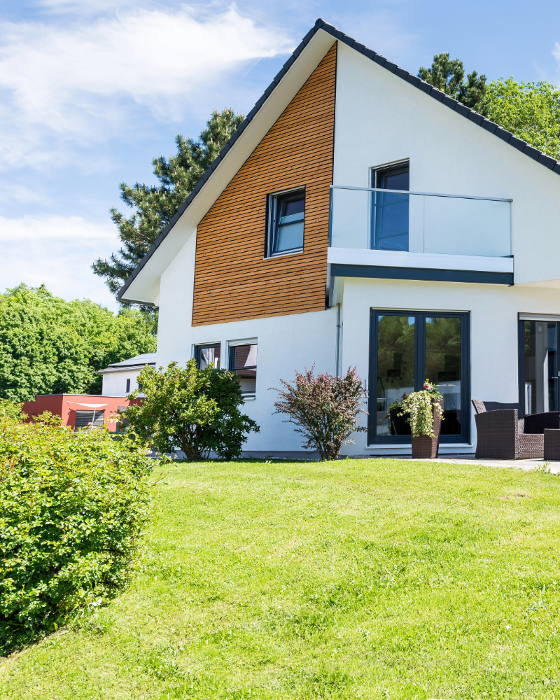 Modern two-story house with white exterior, wooden panel accents, large glass sliding doors, a small balcony, and outdoor seating on a well-maintained lawn surrounded by trees and bushes.