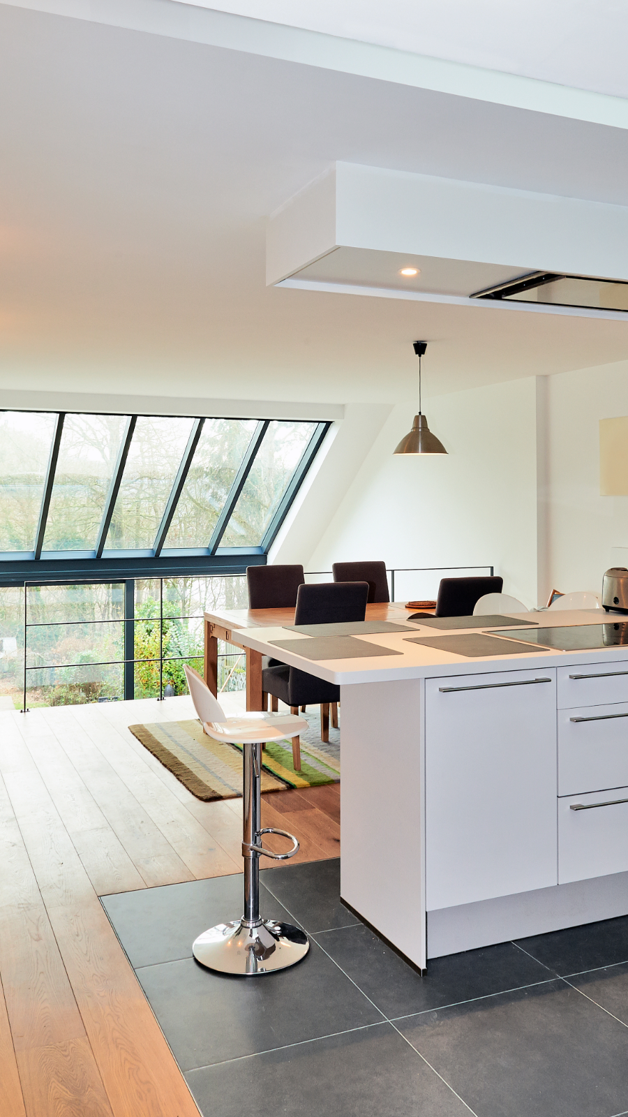 Modern kitchen and dining area with large slanted window, white cabinetry, black chairs around a wooden table, a single white and chrome bar stool, and a hanging pendant light.