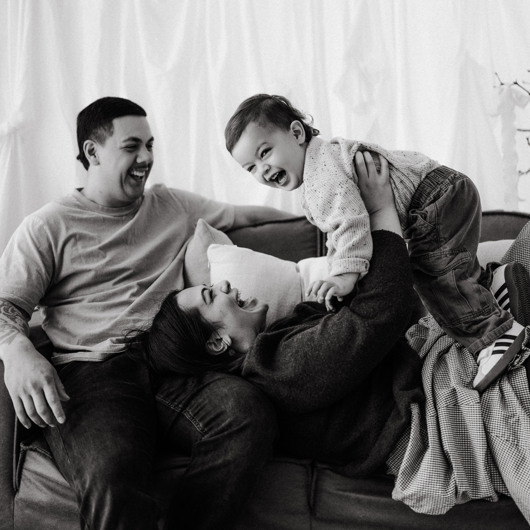 Family playfully wrestling together on a couch, smiling and laughing, in a cozy living room.