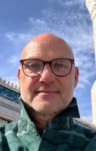 A smiling woman with curly hair wearing glasses, hoop earrings, and layered necklaces, in a cozy indoor setting with blurred background.