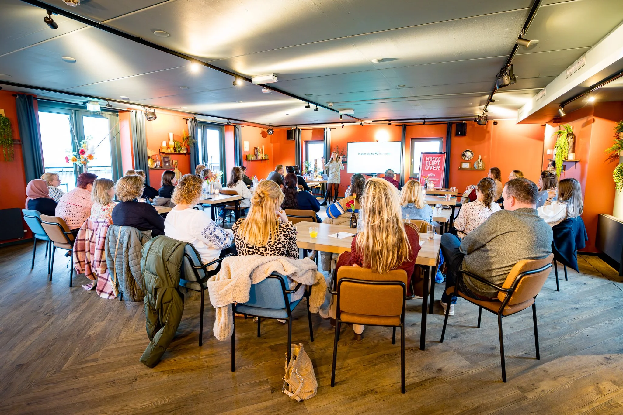 Een groep mensen zit in een vergaderzaal of conferentiehoek en kijkt naar een spreker die voor een scherm staat. De kamer heeft oranje muren, grote ramen met blauwe en witte gordijnen, en is ingericht met planten en decoratieve voorwerpen.