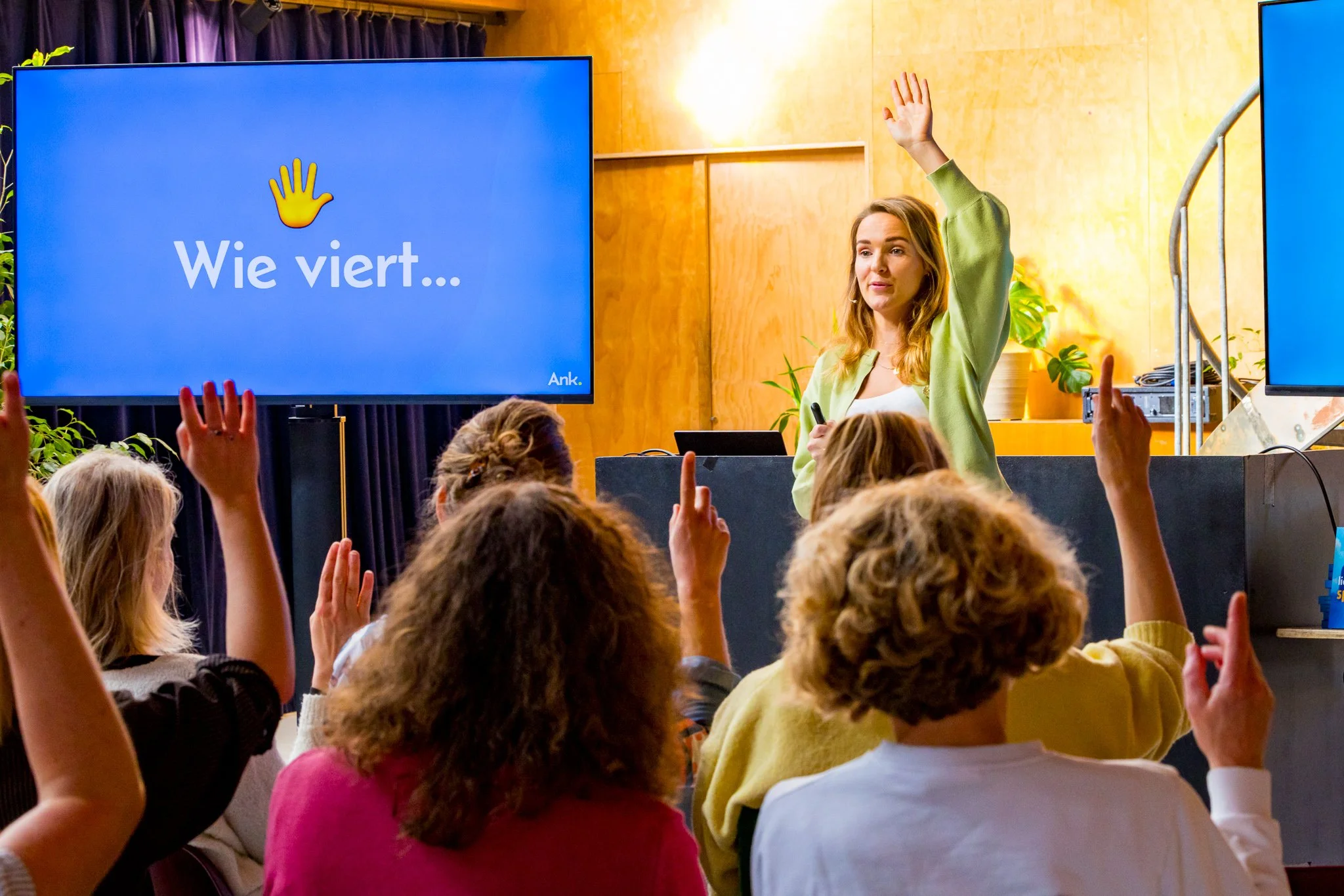 Een vrouw geeft een presentatie voor een groep mensen die vragen met opgestoken handen beantwoorden. Op het scherm staat 'Wie viert...'