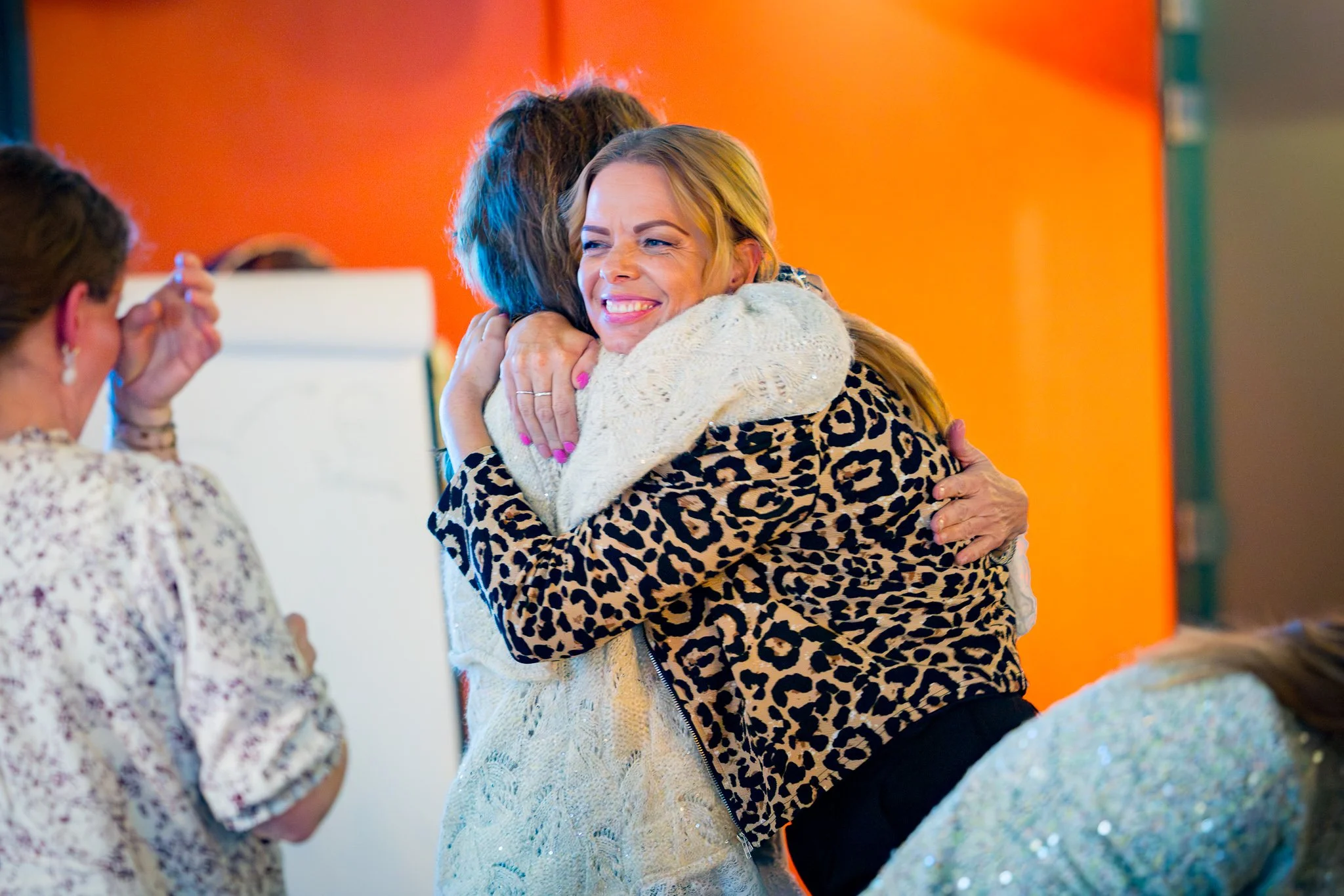 Twee vrouwen omhelzen elkaar blij met een warme hug, terwijl andere mensen eromheen kijken, op een kleurrijke achtergrond.