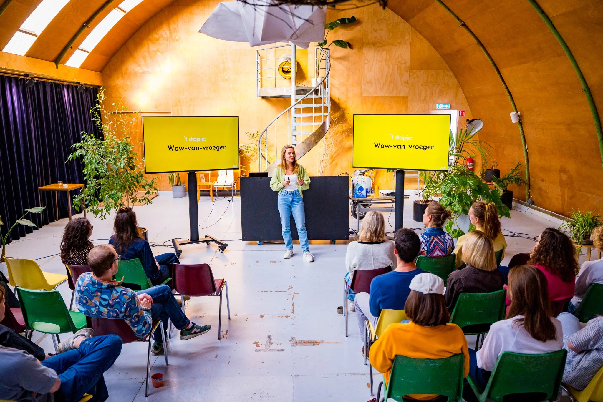Een vrouw geeft een presentatie aan een groep mensen in een kleurrijke, moderne zaal met veel planten, twee grote schermen waarop staat 'Wow-van-vroeger', en een toren van hout en staal.