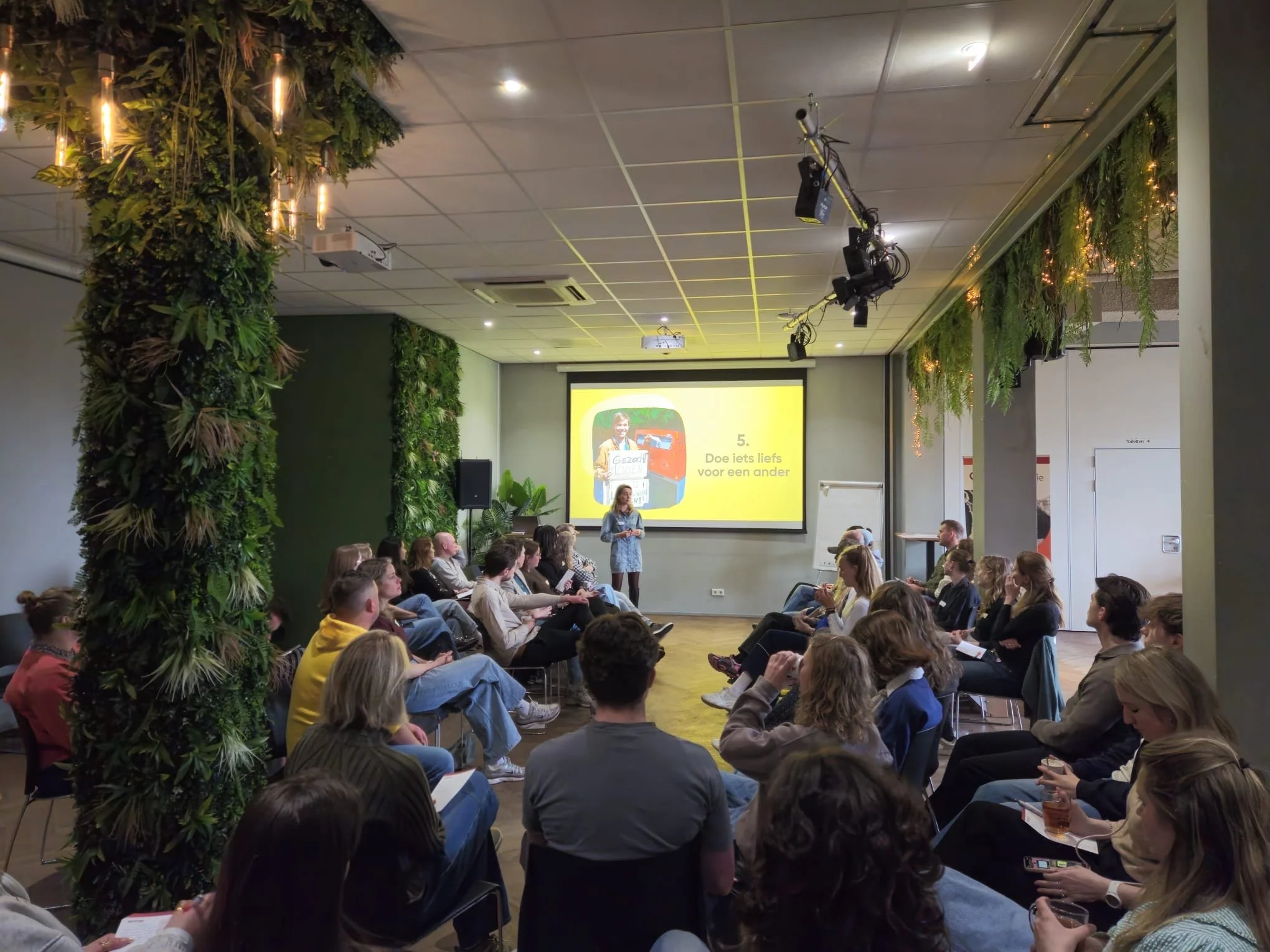 Een zaal vol mensen zit in een halve cirkel voor een spreker die bij een presentatie staat. Op het scherm achter de spreker staat een afbeelding van een persoon met tekst. De zaal is feestelijk versierd met groene planten en lampjes.