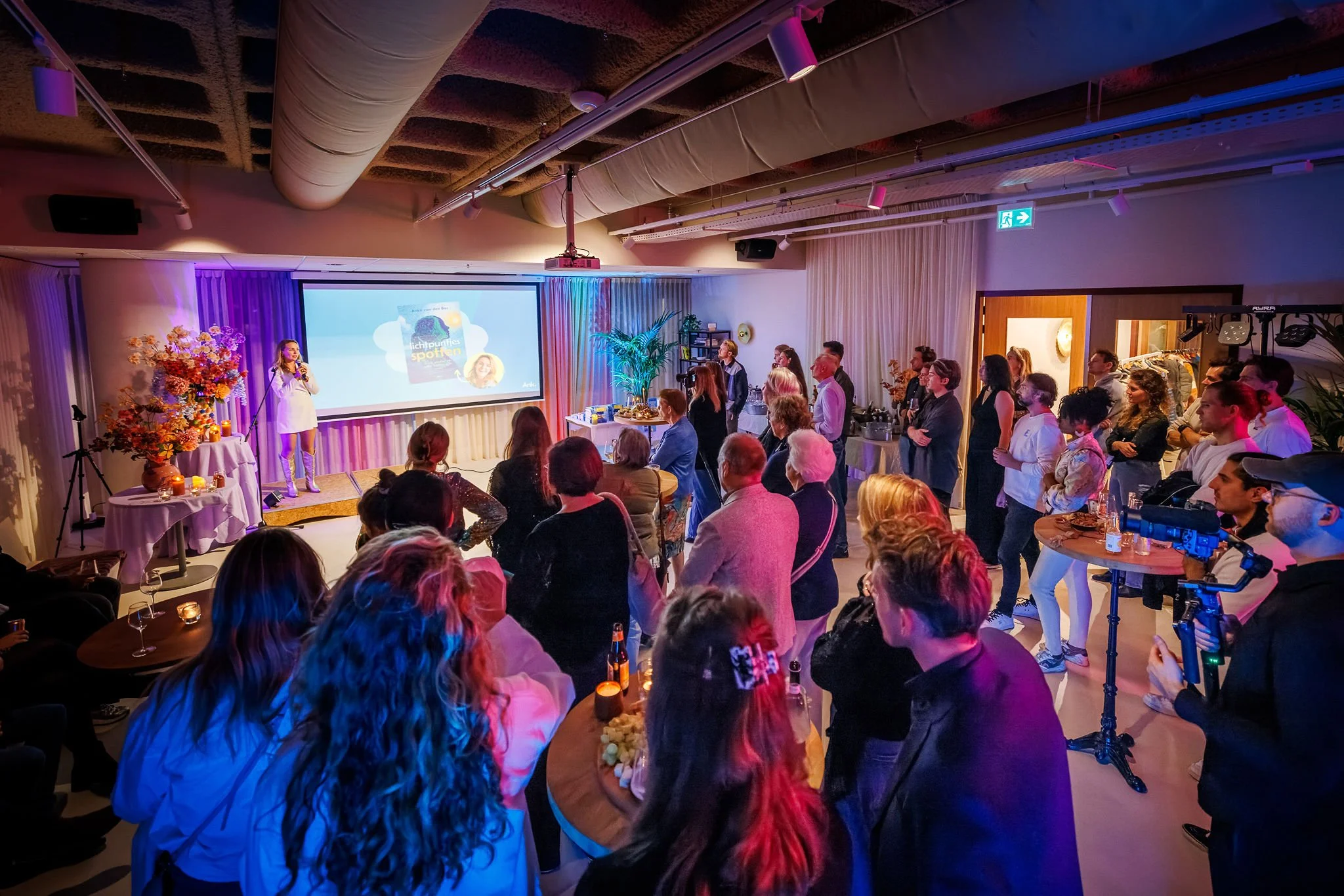 Anke van den Bos staat op een podium met een microfoon voor een publiek. Op de achtergrond een groot scherm en mensen die naar haar luisteren en kijken.
