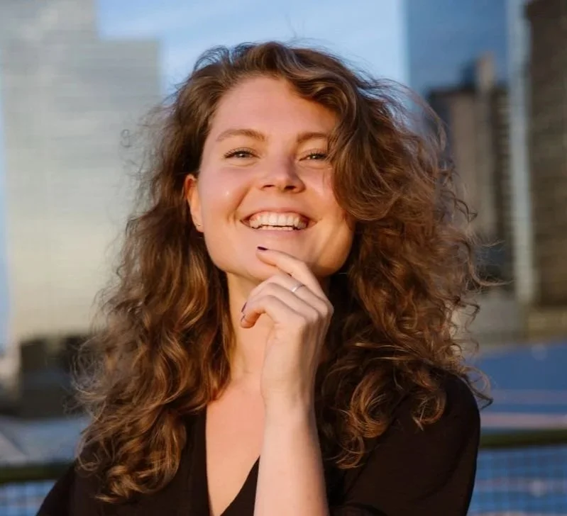 Close-up of a smiling Asian woman with shoulder-length black hair, wearing a blue top, surrounded by green plants in a bright indoor setting.