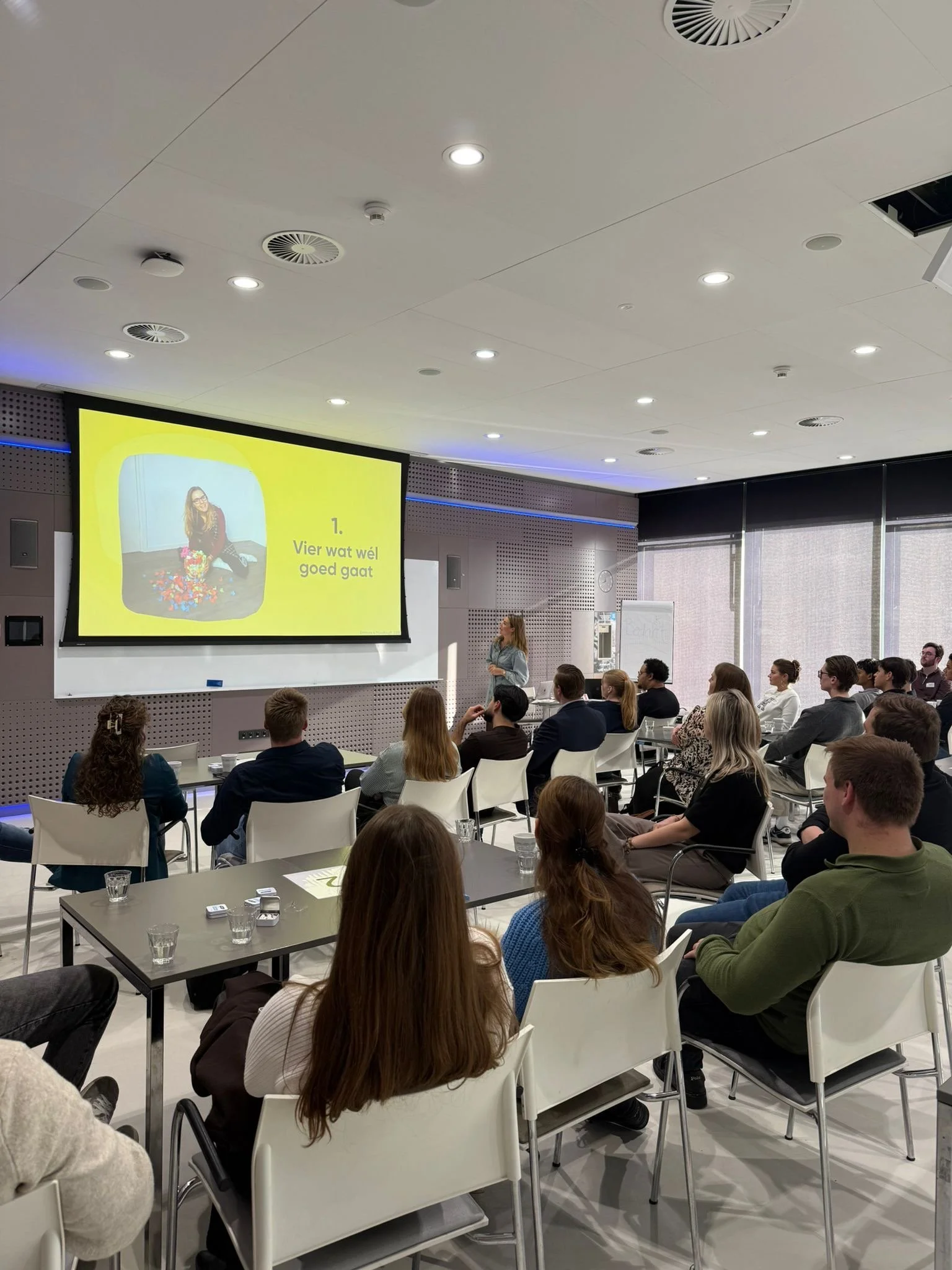 Een presentatie in een vergaderzaal met een grote scherm waarop een dia staat met de tekst "1. Vier wat wél goed gaat" en een afbeelding van een vrouw met bloemen.