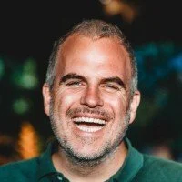 Close-up of a man with curly dark hair, glasses, a nose ring, and a beard, wearing a dark sweater, looking to the side in a bright room.