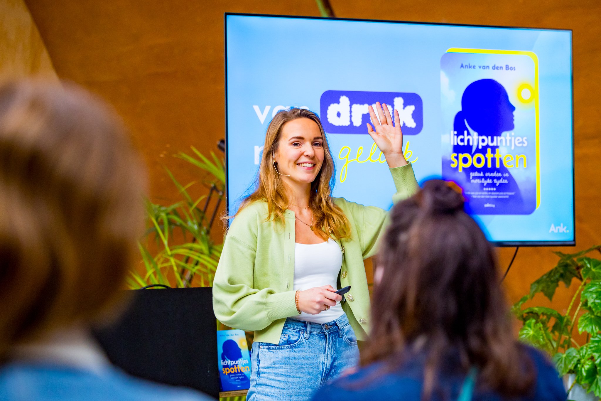 Vrouw geeft een presentatie aan een groep mensen voor een scherm met tekst en afbeeldingen. Ze draagt een lichtgroene blazer en een witte top.