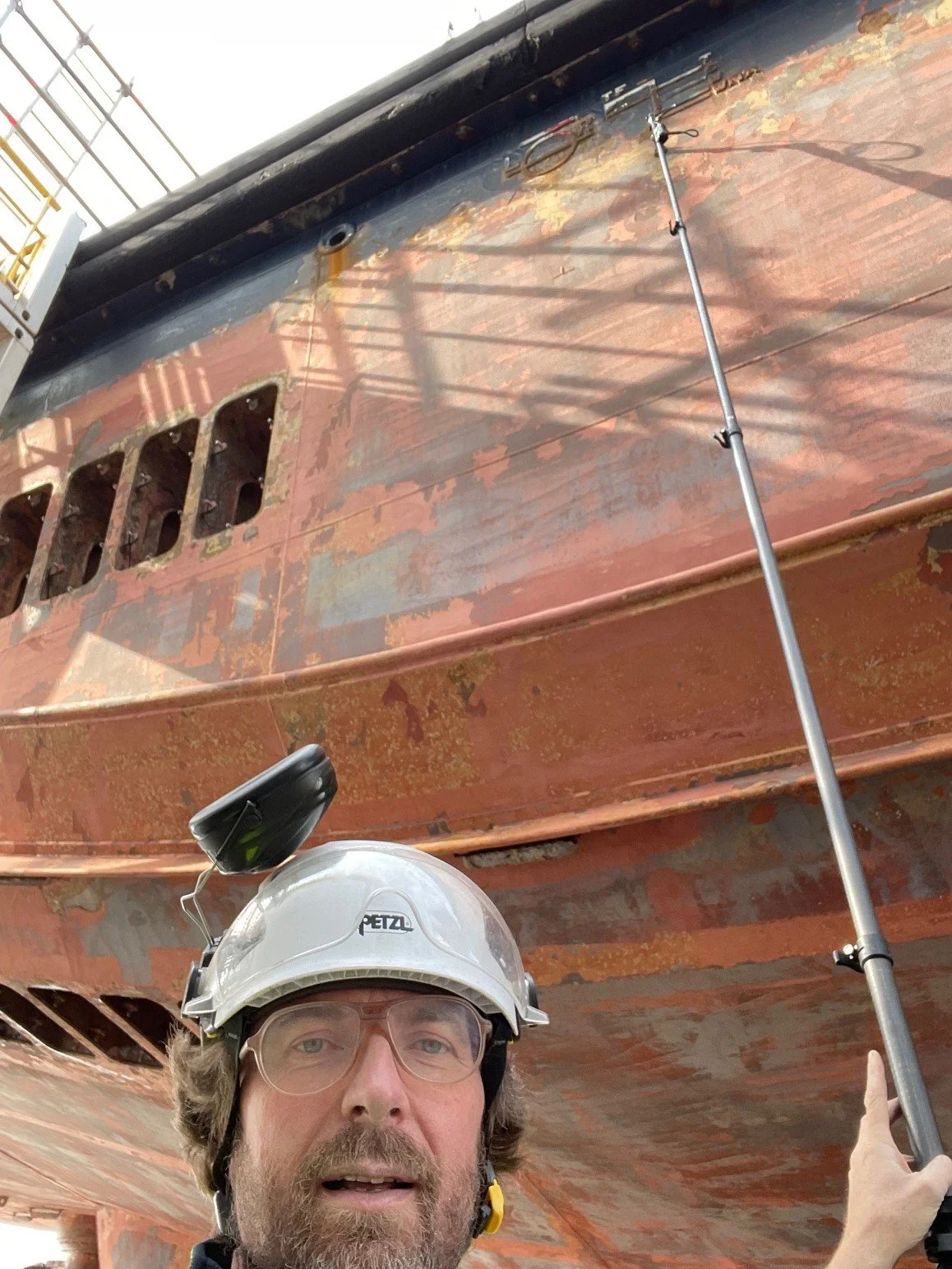 A person wearing a helmet and glasses standing under a large, rusty shipwreck, pointing at it.