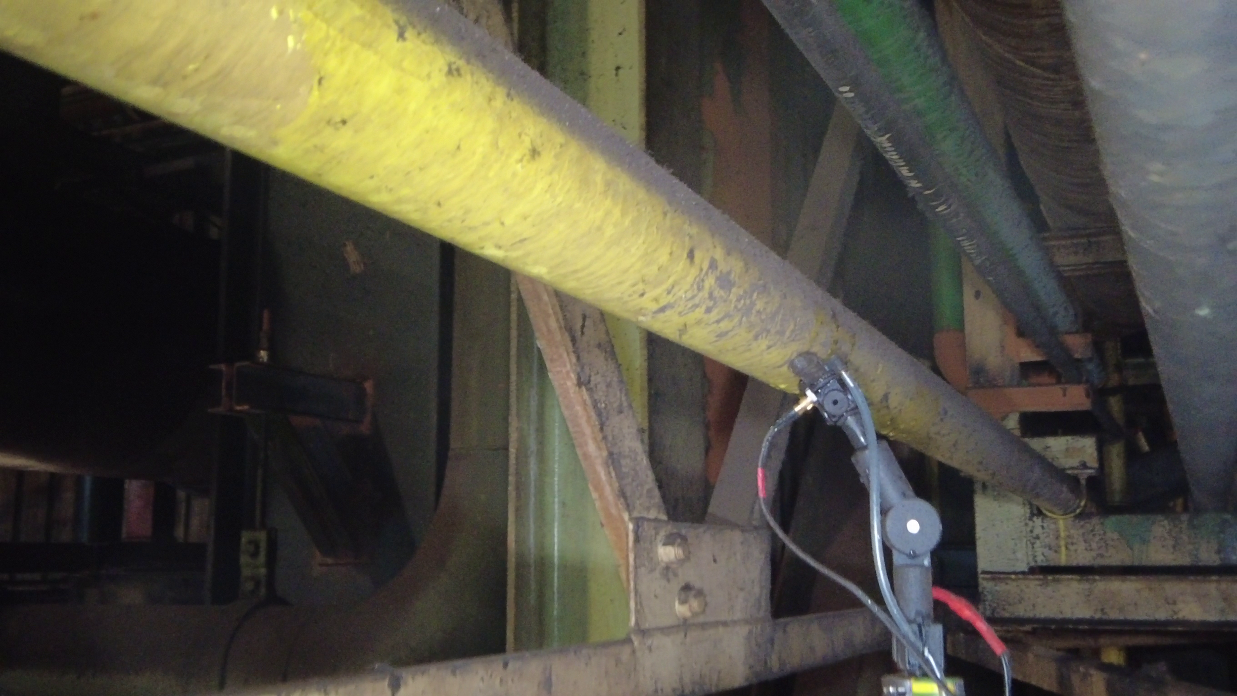 A close-up view of industrial pipes, with a yellow pipe in the foreground and a larger dark pipe to the side, along with some wiring and mechanical components, likely in a factory or mechanical room.