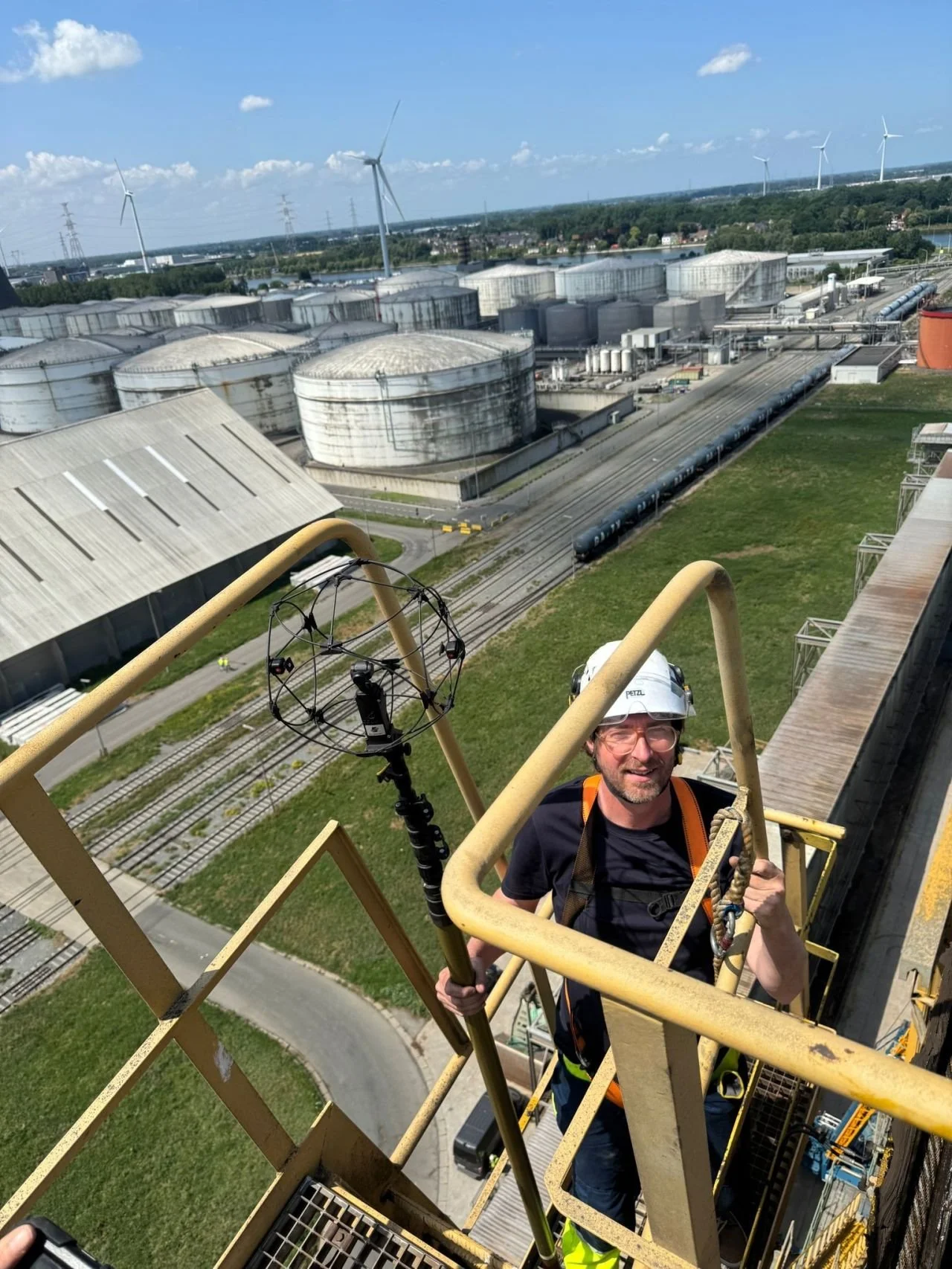 A man wearing a white safety helmet and safety harness stands on a metal platform high above an industrial area with storage tanks, train tracks, and wind turbines in the background on a sunny day.