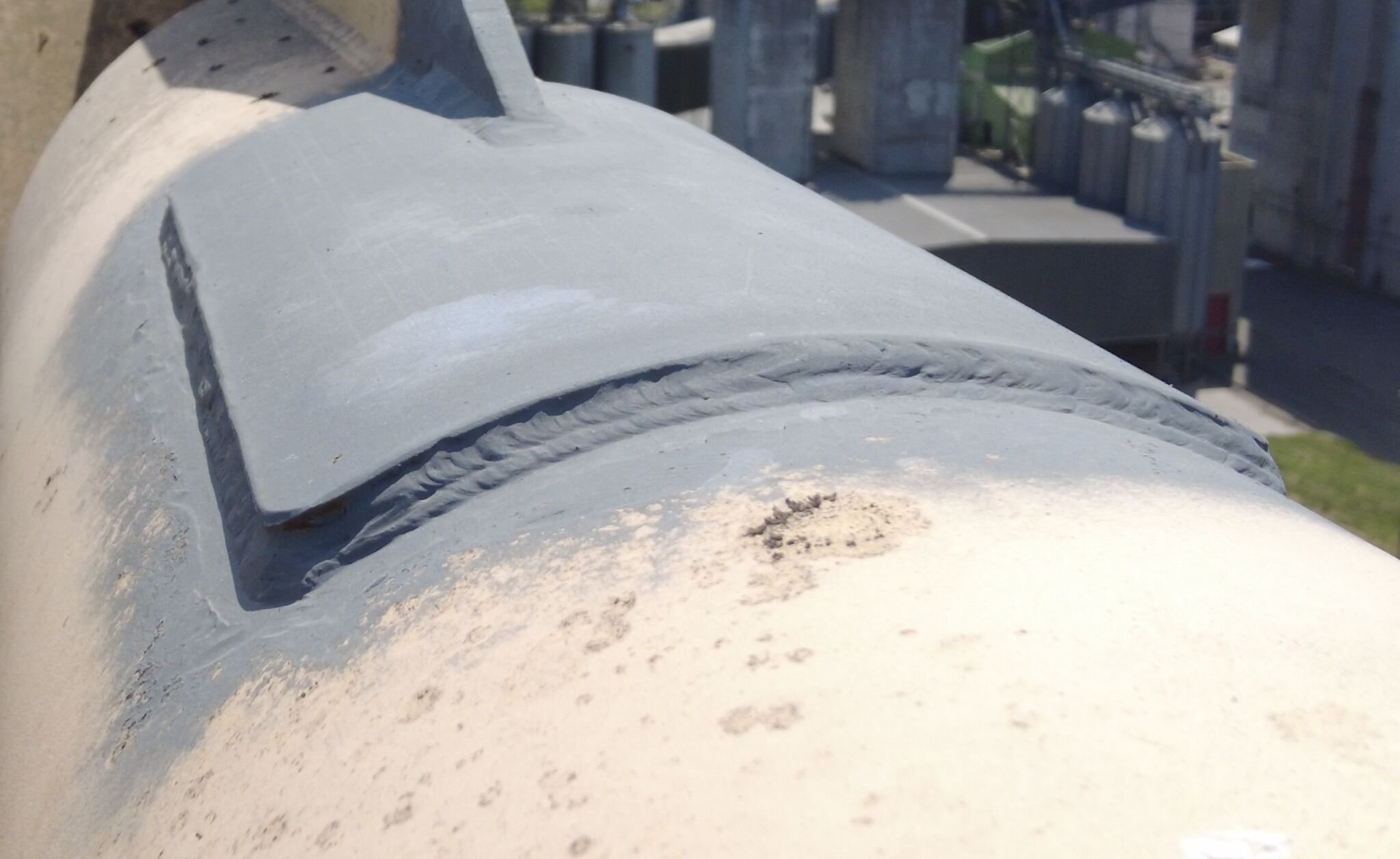 Close-up of a metallic, light gray artillery or telescope mounted on a structure with screws and bolts, overlooking a cityscape with roads and buildings.