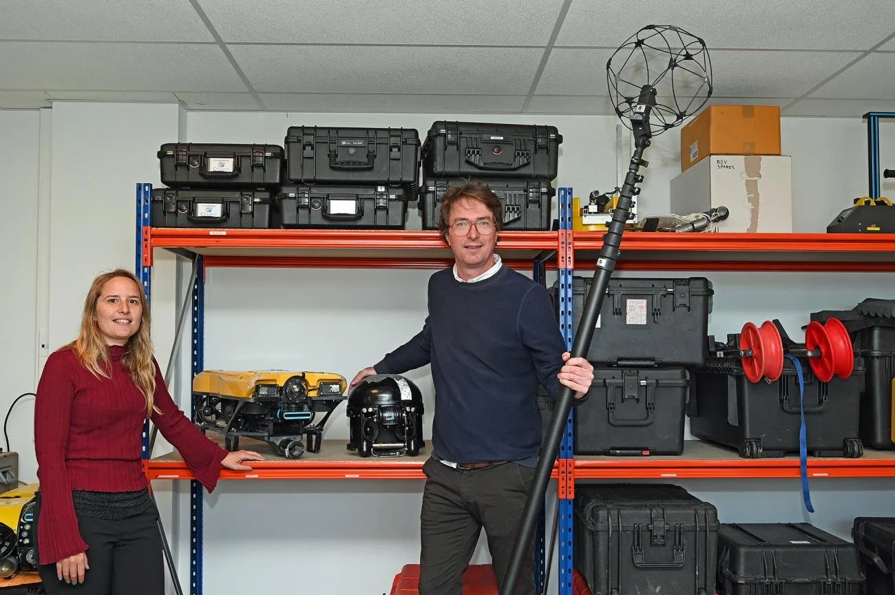 Two people standing in a storage room with shelves containing equipment and tools; one man holding a large drone with a spherical cage on top.