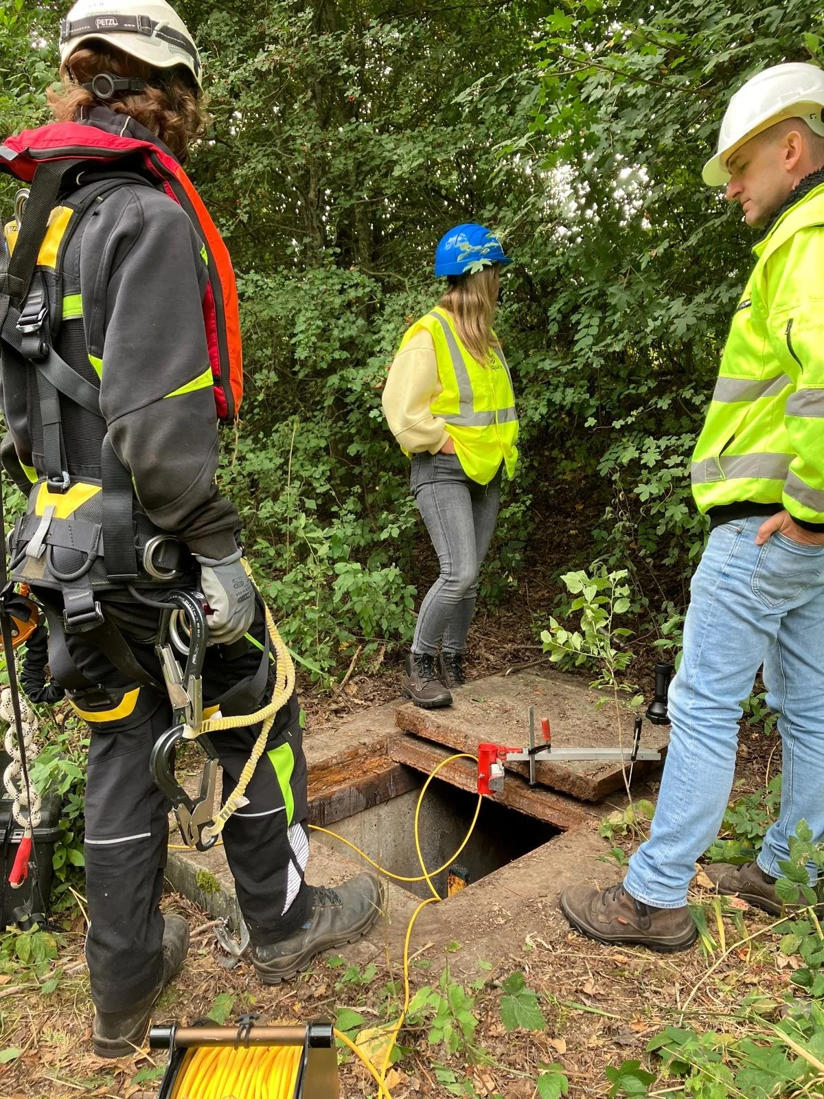 Three workers in safety gear around an open underground utility access, surrounded by greenery.