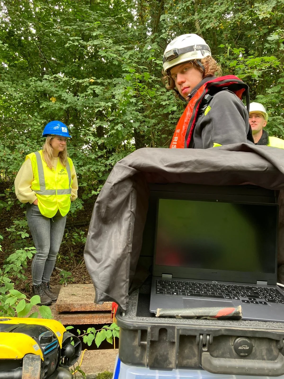 Group of people working outdoors in a forested area with a laptop and safety equipment.