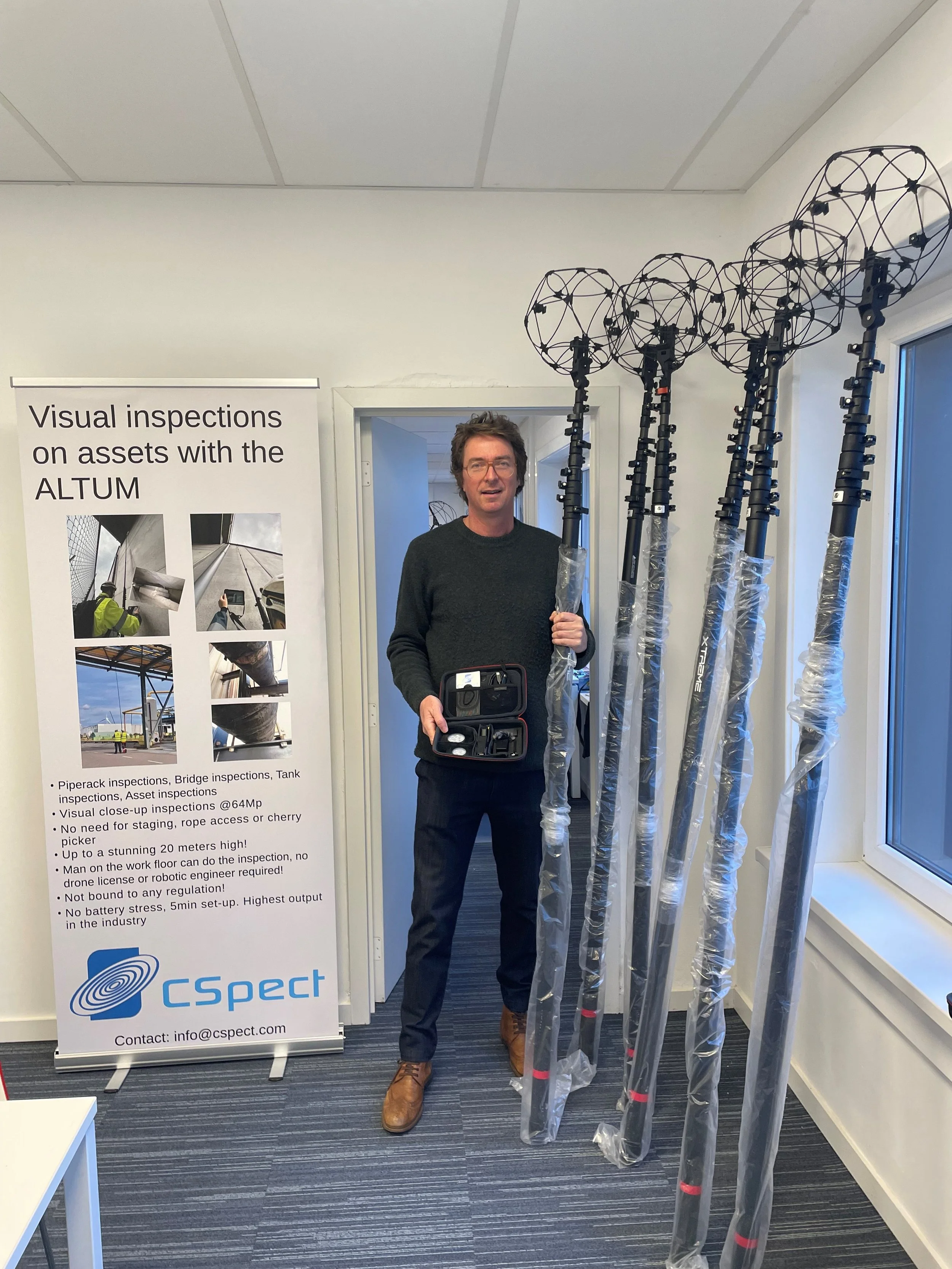 A man standing indoors next to a table of four tall, spiked pole-like antennas and a poster advertising visual inspections of assets with the ALTUM system.