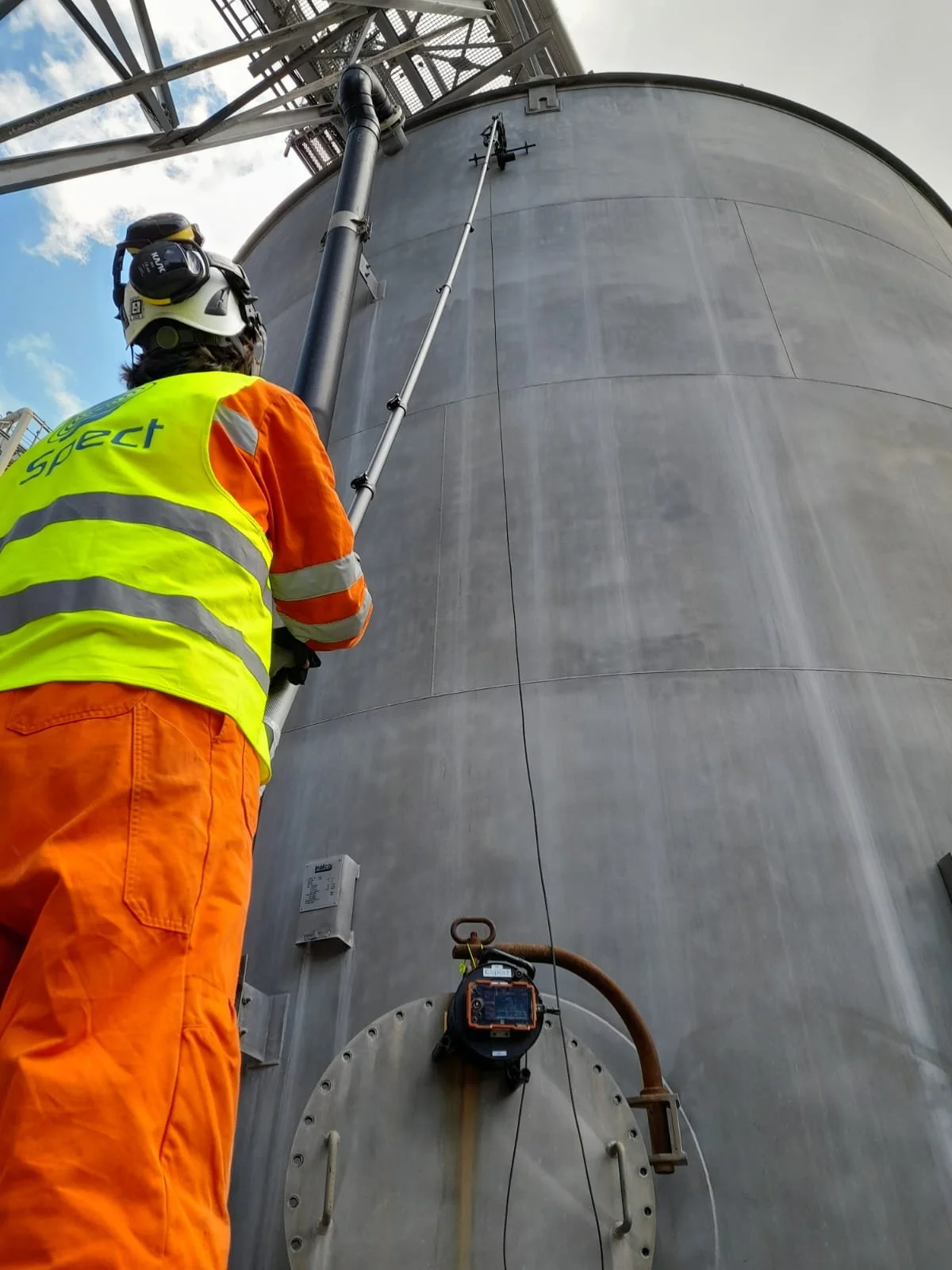 A worker wearing an orange jumpsuit, a yellow safety vest, and a safety helmet with a face shield is inspecting a large industrial storage tank or silo. The worker is looking up and is attached to a long pole or sensor for measuring or inspection purposes, with various pipes and equipment visible on the tank.