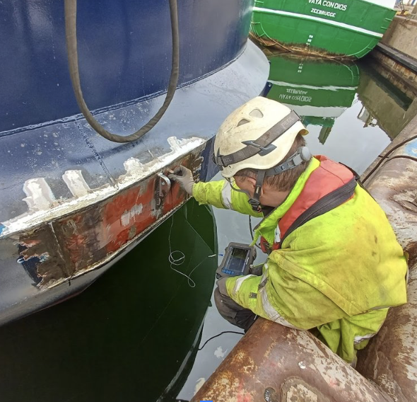 A worker in safety gear inspecting the hull of a large ship near a dock, using tools and a device for measurements.