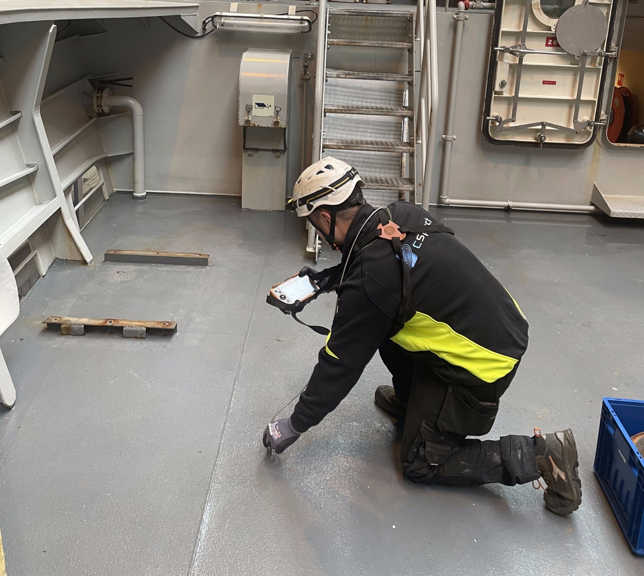 A worker in safety gear kneels on the floor of a vessel or industrial setting, inspecting or working on the floor, with tools and equipment around him.